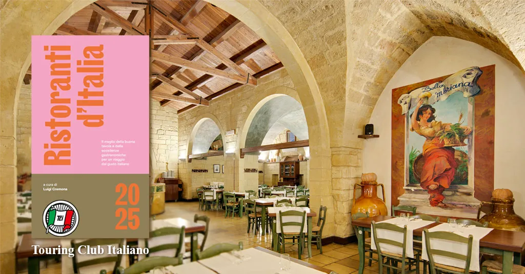 Nell'immagine l'interno della Masseria Bella ‘Mbriana. Ci sono tavoli apparecchiati e sedie verdi. Il muro è a volte con sassi a vista e una parte del soffitto è di legno con sostegni. A sinistra c'è una targhetta posta sopra la foto che dice che la masseria è stata inserita da Touring Club Italiano della guida Ristoranti d'Italia 2025.